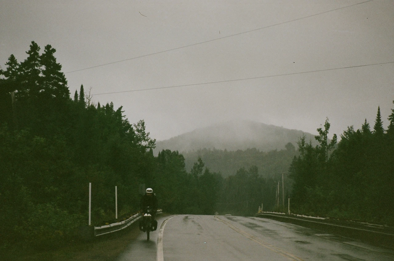 A lone cyclist riding along a wet forest road toward a mountain hidden by rain