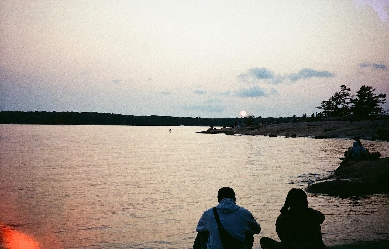 Two anonymous figures sitting beside calm water beneath a low evening sun